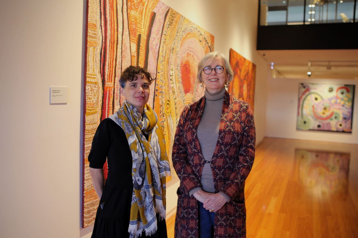 Two women in an art gallery, standing in front of large Aboriginal paintings.